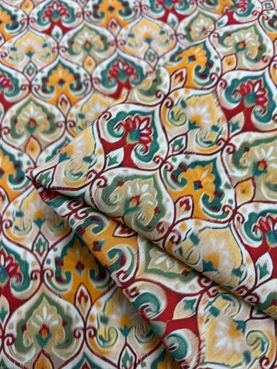 A detailed view of the Designer Rayon - Belle - 145cm from Super Cheap Fabrics highlights a vibrant, intricate repeating floral and geometric pattern in shades of red, green, yellow, and cream. The ornate motifs form a rich tapestry with vivid prints. The fabric's two visible folds make it an adaptable option for various projects.