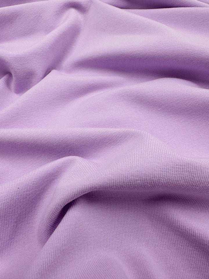 A close-up of Super Cheap Fabrics' Cotton Lycra - Orchid Bouquet reveals a ribbed texture with gentle folds, creating a wavy pattern on the soft, light purple, medium-weight fabric reminiscent of an orchid bouquet.