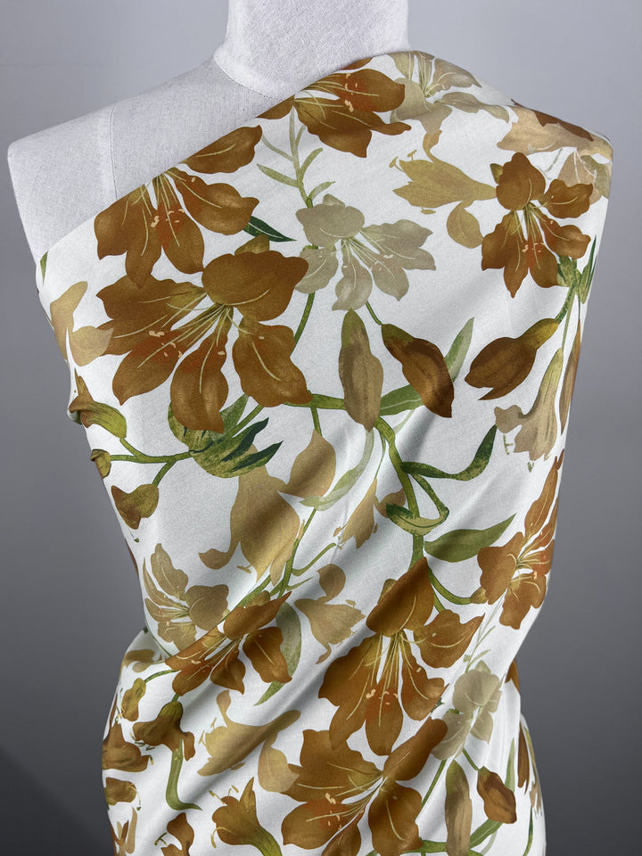 A mannequin is elegantly draped with "Designer Rayon - Autumn Gold - 145cm" from Super Cheap Fabrics, featuring a botanical pattern of stylized brown and green leaves and flowers set against a white background. The vibrant print offers a bold and organic look, creating a natural and sophisticated aesthetic that adds versatility to any wardrobe.