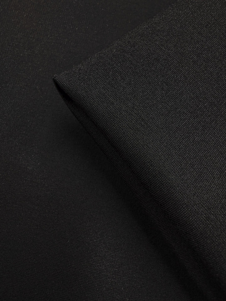 Close-up of Milano Ponte - Black - 160cm by Super Cheap Fabrics, highlighting its smooth texture and subtle weave. A folded corner creates a sharp edge, showcasing this versatile heavyweight fabric's quality.