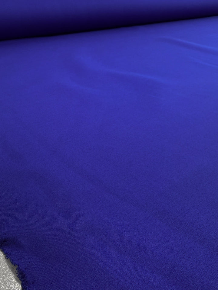 A close-up of Super Cheap Fabrics' Milano Ponte - Dazzling Blue (160cm) shows its smooth, vibrant surface with soft folds and a slightly frayed edge in the lower left corner.