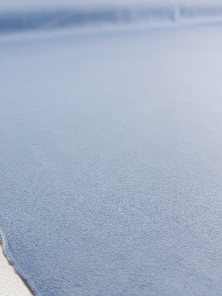 A close-up of Super Cheap Fabrics' Wool Cashmere - Cool Blue (152cm) shows light blue textured paper with gentle lighting highlighting its soft, luxurious surface and color variation, evoking the feel of wool cashmere.