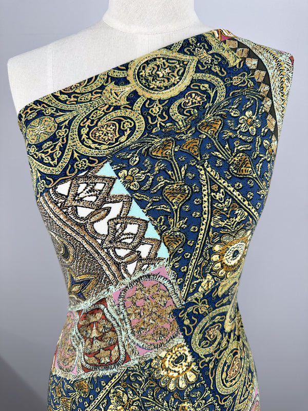 A close-up of an elegant one-shoulder dress on a mannequin, made from medium-weight polyester/spandex fabric. The tapestry pattern features gold, navy blue, and pastel in geometric and floral designs, crafted from Super Cheap Fabrics' Printed Lycra - Mosaic Patchwork - 150cm.