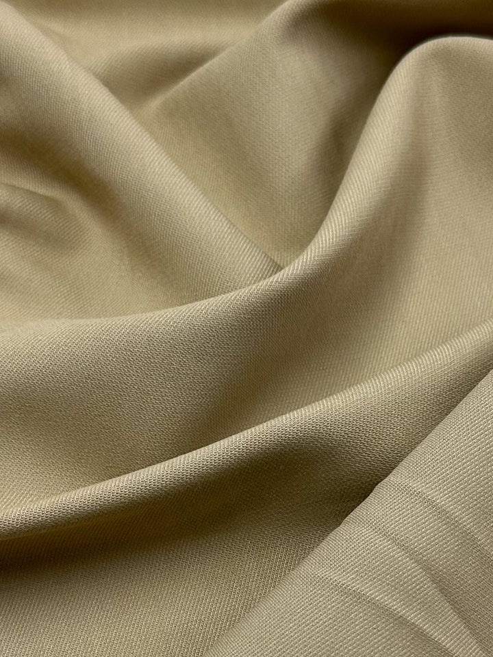 A close-up of the Stretch Viscose Suiting fabric in Pebble by Super Cheap Fabrics showcases its smooth, slightly glossy texture with gentle folds. Its soft, beige twill and elegant fluidity make it ideal for sophisticated suiting garments. Width: 130cm.