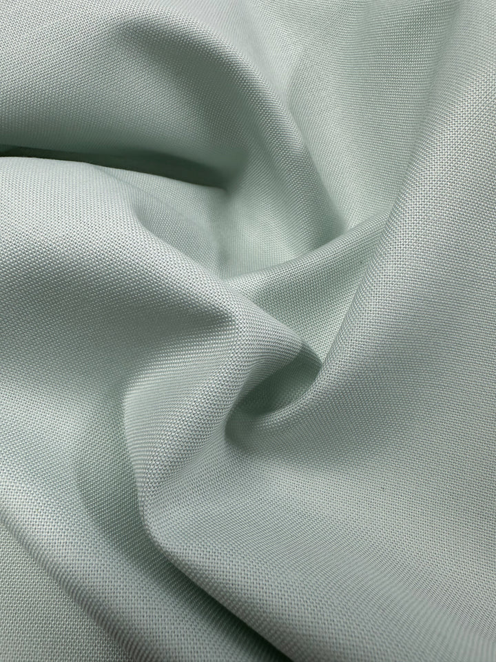 Close-up of "Linen Blend - Blue Flower - 140cm" by Super Cheap Fabrics, showcasing a light green cotton linen blend fabric with a subtle weave pattern. It features slight wrinkles that create gentle folds and shadows to highlight its texture.