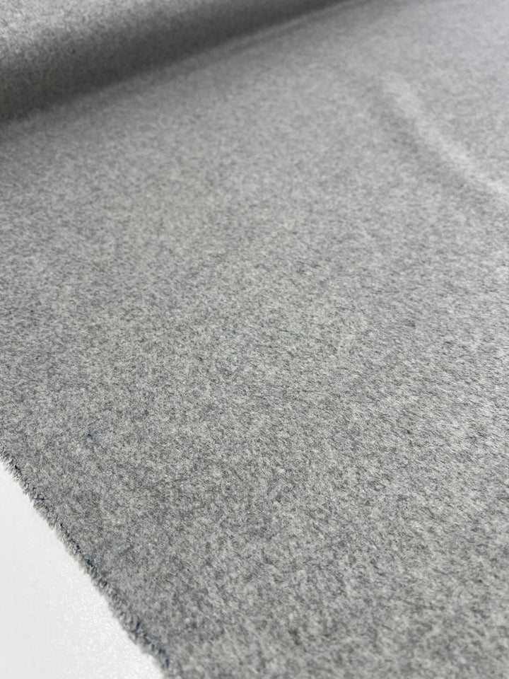 A close-up view of the Wool Cashmere - Grey Marle - 150cm from Super Cheap Fabrics shows a flat, light gray fabric with a soft texture. The extra heavy weight wool cashmere is laid out smoothly with the edge slightly visible in the lower part of the image, displaying a subtle weave and consistent color. Dry clean only to maintain its quality.