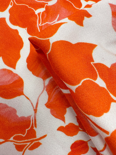 Close-up of Super Cheap Fabrics’ Mercerised Rayon - Flameleaf (150cm): breathable, lightweight rayon with a bold orange floral pattern on white, soft-edged flowers, subtle pink highlights, and a visible fabric fold.
