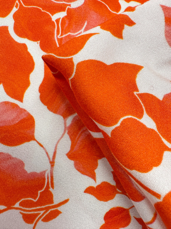 Close-up of Super Cheap Fabrics’ Mercerised Rayon - Flameleaf (150cm): breathable, lightweight rayon with a bold orange floral pattern on white, soft-edged flowers, subtle pink highlights, and a visible fabric fold.