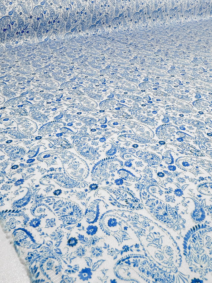 Super Cheap Fabrics’ Mercerised Rayon - Delft Vine (150cm) features a blue and white paisley pattern with intricate floral and leaf designs, silky soft to the touch, seen spread flat across a surface and into the background.