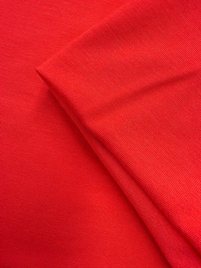 A close-up of Super Cheap Fabrics' Cotton Lycra - Hibiscus (165cm) shows the smooth, bright red stretchy fabric with a folded corner, highlighting its texture and weave.