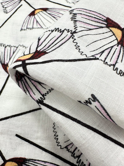 The Linen Blend - Lily from Super Cheap Fabrics is a white fabric featuring stylized pink and yellow floral patterns outlined in black. Perfect for lightweight children's clothing, the 147cm wide material appears slightly folded, highlighting its texture and design.