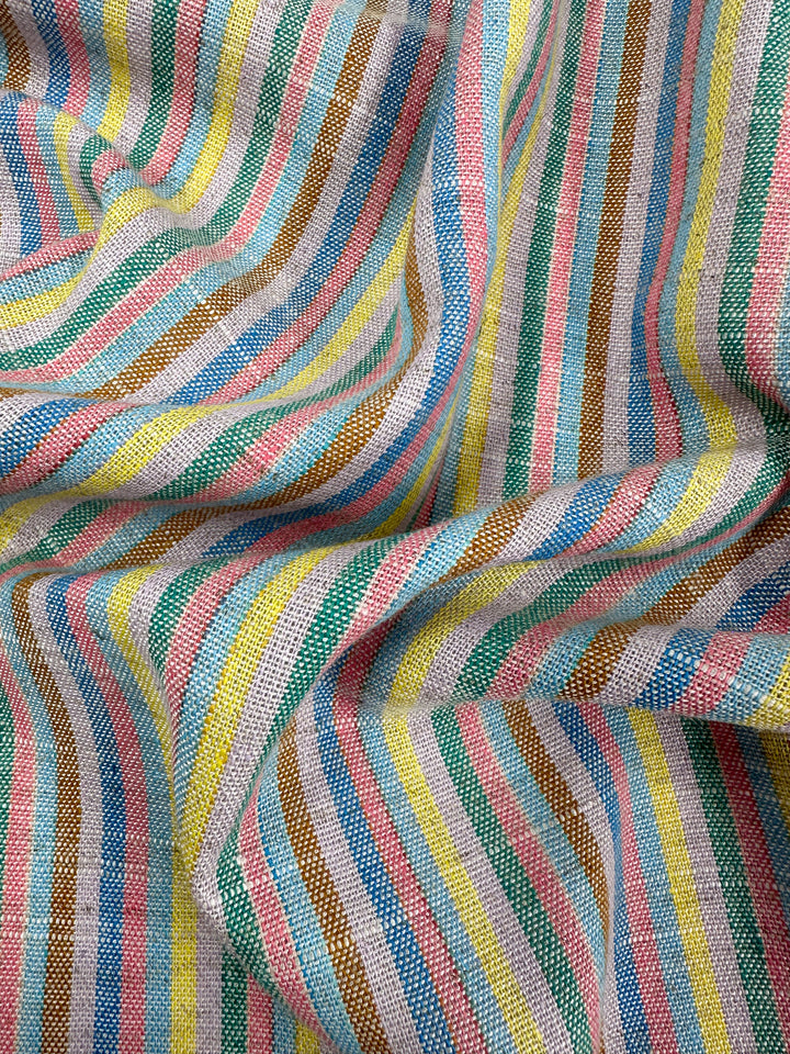 The Linen Blend - Tropicana - 143cm from Super Cheap Fabrics is a yarn-dyed fabric with vertical stripes in blue, pink, yellow, green, and brown. It features visible weaves and a gently draped or wrinkled appearance, giving it a charming vintage look.