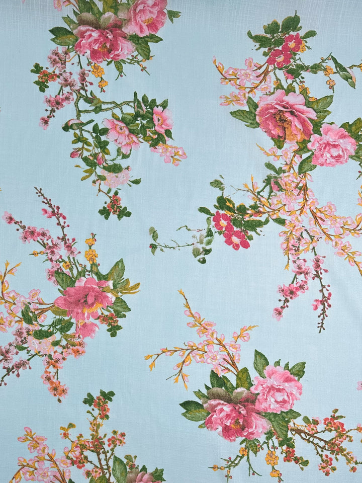 Super Cheap Fabrics' Linen Blend - Pastel Garden (147cm) features a floral pattern with pink roses, green leaves, and small orange and pink blossoms on light blue linen. The pastel colors are evenly distributed for a gentle and elegant look with a lightweight feel.