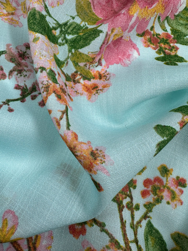 A close-up of the Linen Blend - Pastel Garden - 147cm by Super Cheap Fabrics reveals a floral design with pink and orange flowers and green leaves against a light blue background. Known for pastel colors, this fabric has a soft texture and appears slightly draped.