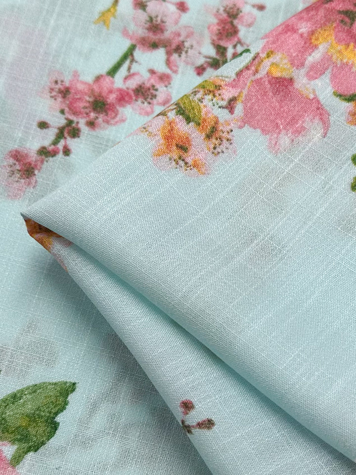 Close-up of Super Cheap Fabrics' Linen Blend - Pastel Garden, 147cm wide. The light blue fabric features a floral print with pink and orange flowers and green leaves. Its lightweight texture is beautifully layered and folded, showcasing its pastel colors.