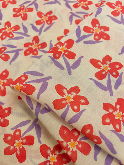 The Linen Voile - Bloom - 150cm by Super Cheap Fabrics features red flowers with purple stems printed on a beige, lightweight voile fabric, beautifully showcasing an elegant fold.