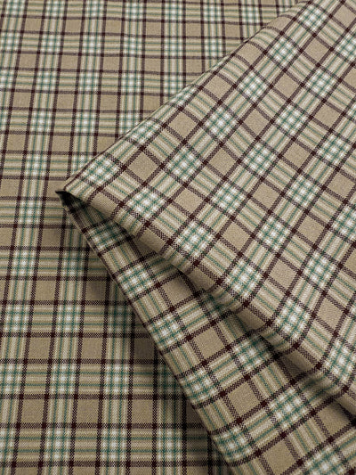 A folded view of the Shirting - Safari fabric by Super Cheap Fabrics, showing its beige and green plaid pattern on a complementary background. The design includes intersecting green, brown, and white lines crafted from a polyester cotton blend. This light to medium weight fabric is ideal for children's clothing due to its timeless tartan style.