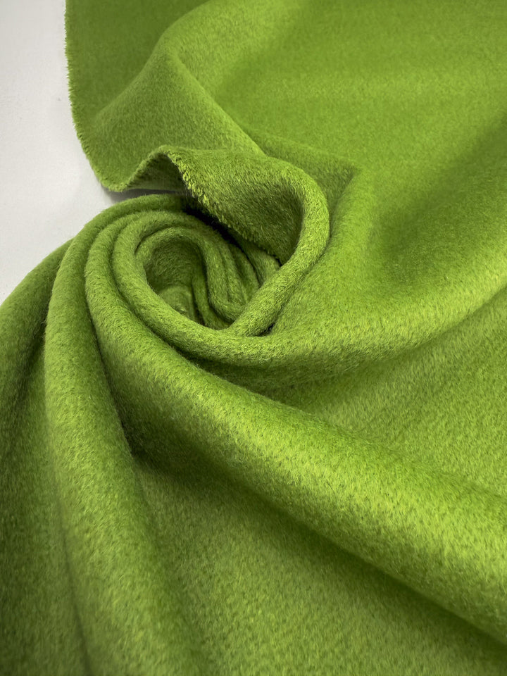 A close-up of Wool Cashmere - Titanite (154cm) by Super Cheap Fabrics showing its bright green, soft, fuzzy luxury texture, loosely rolled into a spiral on a white surface.