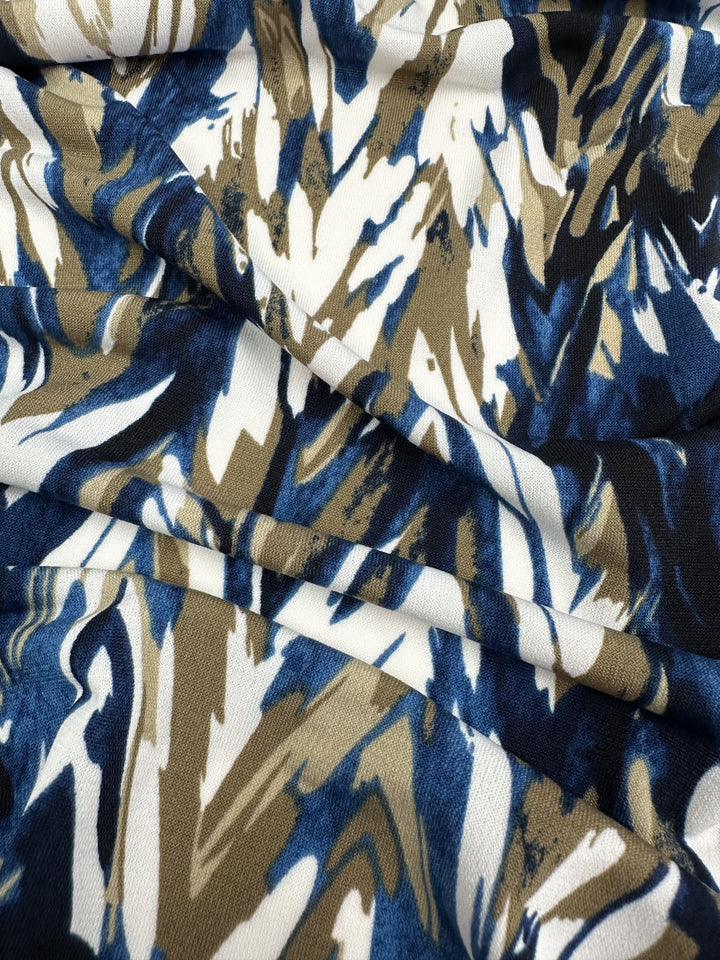 A close-up of Super Cheap Fabrics' Printed Lycra - Arrows - 150cm showcases a medium-weight fabric with a camouflage-like pattern in blue, brown, and white. The Polyester/Spandex blend appears slightly crumpled, highlighting its texture and color mix.