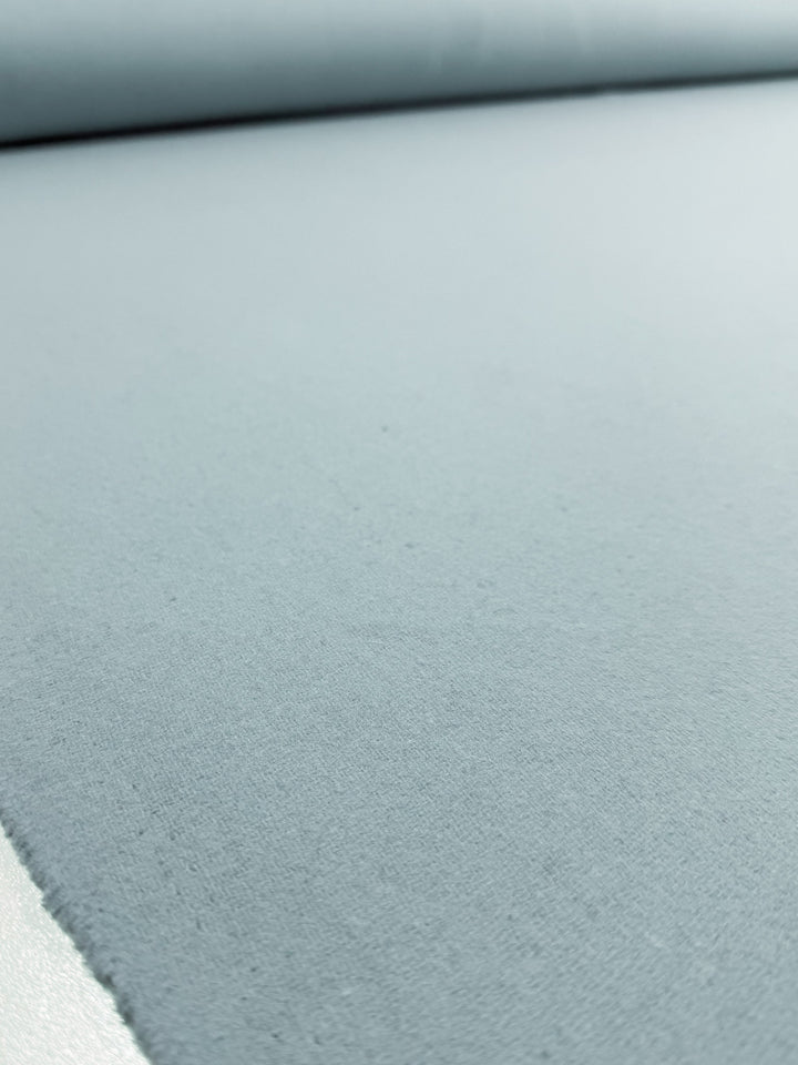 A close-up of Super Cheap Fabrics' Wool Cashmere - Blue Glass (150cm), showing the smooth, luxurious blue fabric laid flat with a matching roll in the background.