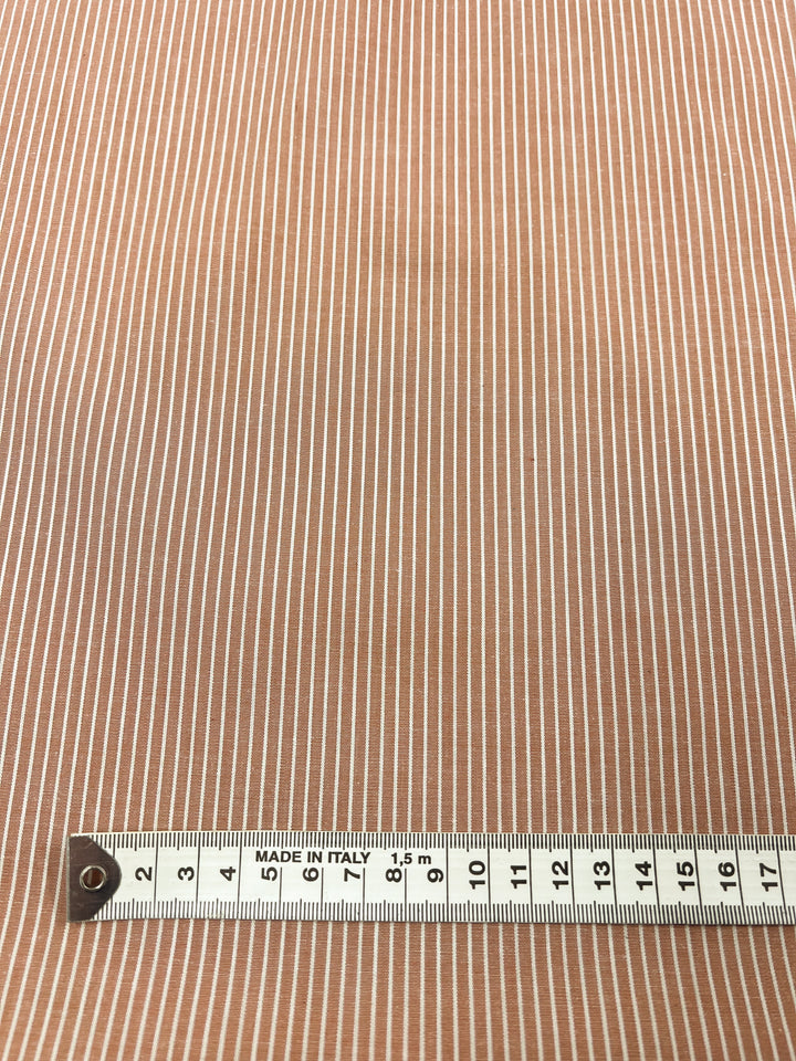 A detailed view showcases the lightweight, brown "Shirting - Marmalade - 147cm" polyester cotton fabric from Super Cheap Fabrics, featuring thin white vertical stripes. Positioned horizontally along the bottom edge is a measuring tape marked "Made in Italy," displaying measurements in centimeters up to 19.