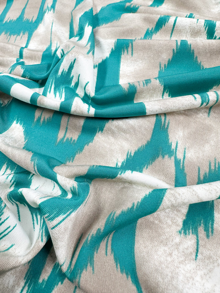 A close-up of Super Cheap Fabrics' Printed Lycra - Dynasty shows a medium-weight fabric with a teal and white zigzag pattern on beige, where draped folds create shadows enhancing its texture.