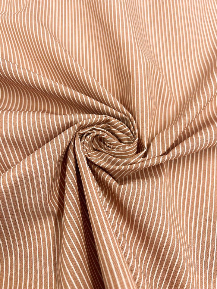 Close-up of light brown, lightweight Shirting - Marmalade - 147cm by Super Cheap Fabrics, featuring fine white vertical stripes intricately gathered into a spiral pattern at the center, creating folds and texture reminiscent of marmalade swirls.