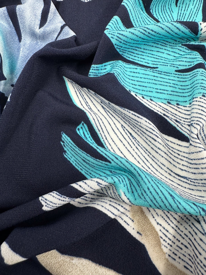 Close-up of medium-weight Printed Lycra fabric with a bold leaf pattern in teal, cream, and navy. The textured surface and fine outlines provide an abstract, dynamic design ideal for those seeking durability and vibrant style. Product: Frost Tropic - 150cm by Super Cheap Fabrics.