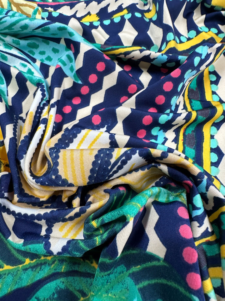 A close-up of colorful Printed Lycra fabric by Super Cheap Fabrics, featuring an abstract design with zigzags, dots, and geometric shapes in blue, green, pink, yellow, and white. The medium-weight Palm Springs fabric gently gathers into folds and contours. Width: 150cm.