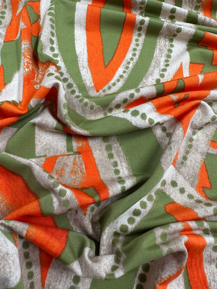 The Printed Lycra - Papaya by Super Cheap Fabrics features an abstract design with orange, green, and white circles and triangles. It measures 150cm wide and has a smooth texture, neatly gathered in folds.