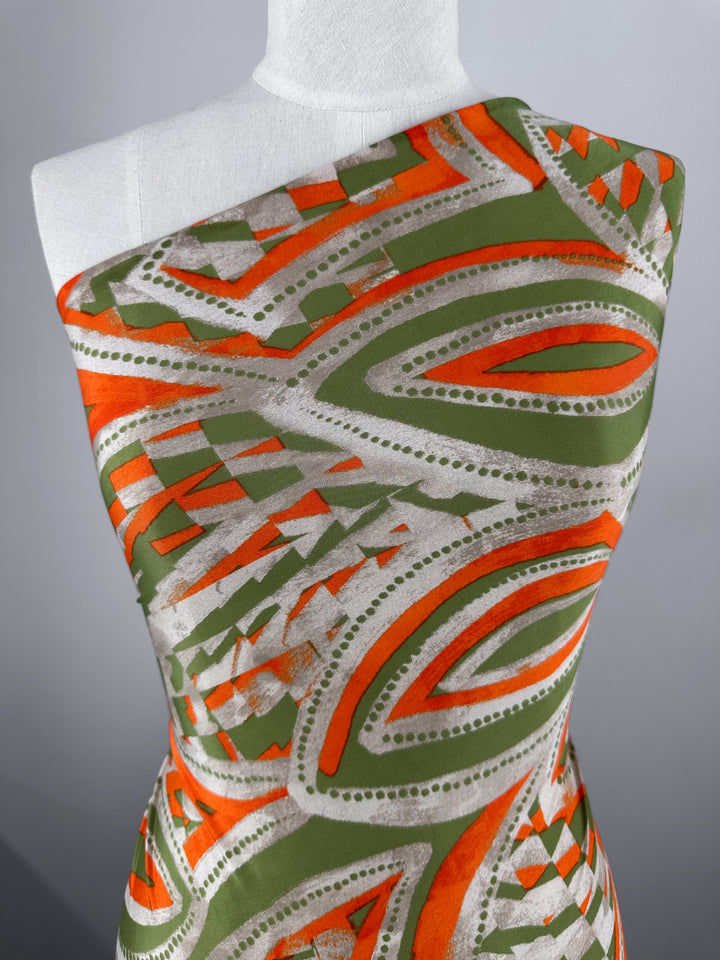 A mannequin draped in Super Cheap Fabrics' Printed Lycra - Papaya, a medium-weight fabric with bold geometric patterns in orange, green, and silver. The design features dynamic curved and angular shapes for a colorful visual effect.