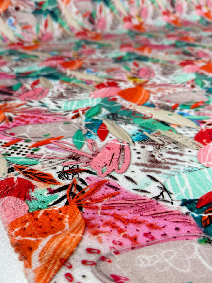 A close-up of Mercerised Rayon - Electric Orchard (148cm) by Super Cheap Fabrics shows vibrant abstract shapes in pink, red, orange, green, and teal with floral and botanical motifs, highlighting the fabric's colorful texture and striking design.