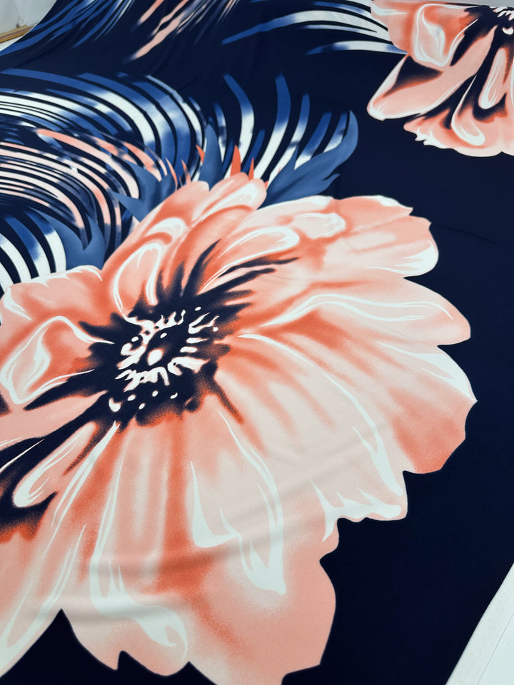 A close-up view of Super Cheap Fabrics' Printed Lycra - Bold - 150cm shows medium-weight fabric with large floral patterns in peach, white, and dark blue. The design highlights prominent blooms with intricate petal details against a contrasting dark background.