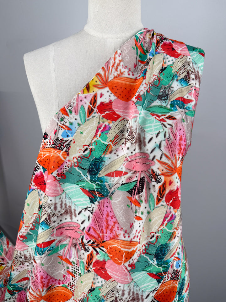 A mannequin displays Super Cheap Fabrics’ Mercerised Rayon - Electric Orchard (148cm), a silky soft fabric with a vibrant abstract floral pattern in red, pink, teal, green, and white, set against a plain gray background.