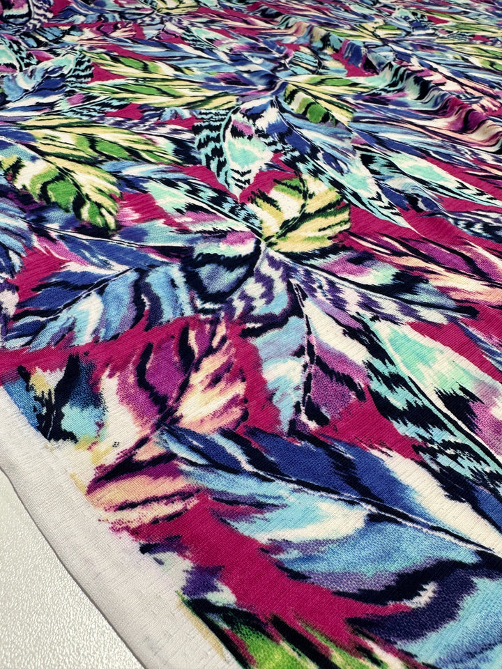 The Super Cheap Fabrics' Printed Lycra - Wavy (150cm) is a vibrant medium-weight fabric with an abstract pattern in lively shades of pink, blue, green, and yellow, resembling artistic brushstrokes or feathers on its Polyester/Spandex surface.