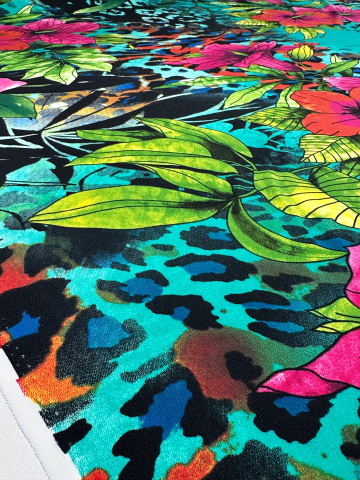 The "Printed Lycra - Aqua Flora - 150cm" by Super Cheap Fabrics is a vibrant medium-weight fabric featuring tropical leaves and flowers in green, pink, and red over a teal and black leopard print. Made from durable Polyester/Spandex for added stretch.