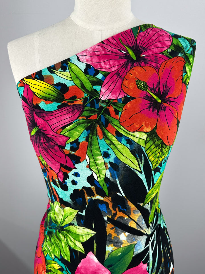 A medium-weight polyester/spandex dress displayed on a mannequin features Super Cheap Fabrics' Printed Lycra - Aqua Flora, showcasing large pink and orange flowers, green leaves, and abstract patterns on a black background.