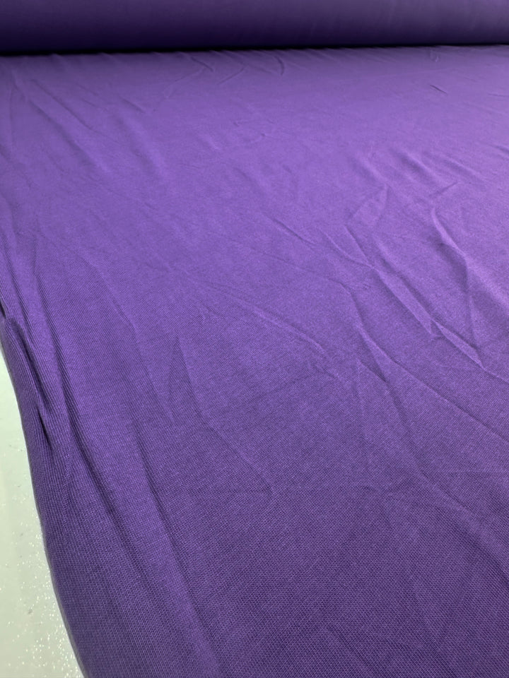 A large piece of the Super Cheap Fabrics' Cotton Jersey in Patrician Purple, measuring 170cm and medium weight, is spread out, showcasing its smooth texture with natural folds and wrinkles.