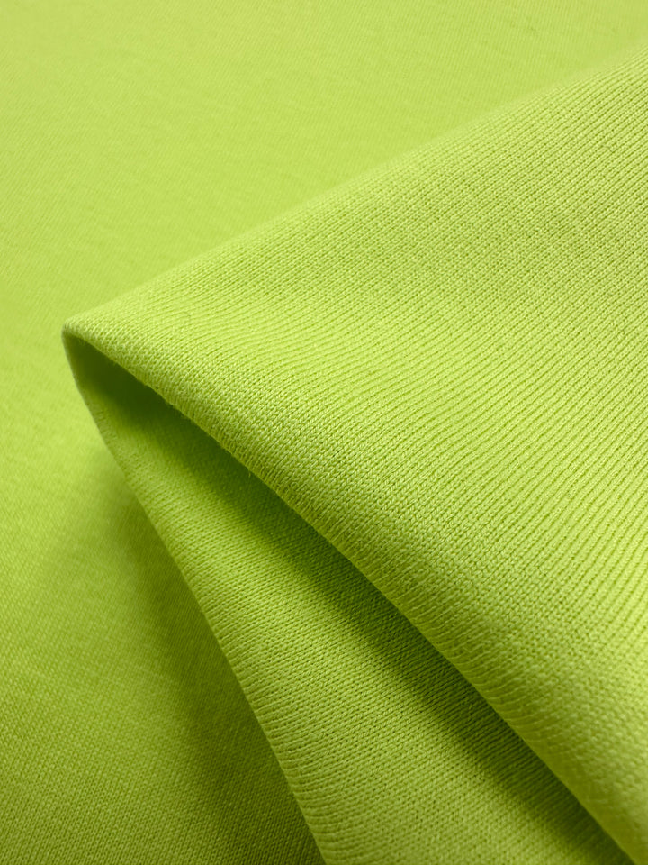 A close-up of the Super Cheap Fabrics Cotton Jersey in Daiquiri Green shows a folded edge with a smooth knit texture and subtle vertical lines, enveloping the image in vibrant color. Width is 170cm.