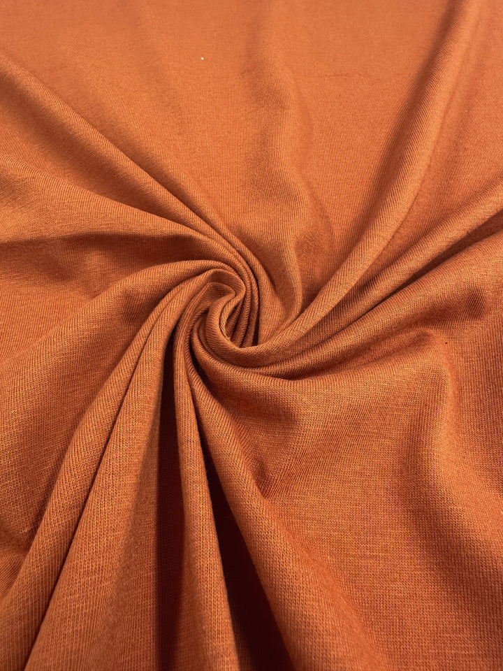 A close-up of Super Cheap Fabrics' Cotton Jersey in Apricot Orange, 160cm wide, shows the fabric draped in a spiral pattern with dynamic folds that highlight its soft, smooth texture and emphasize its flexibility and warmth.