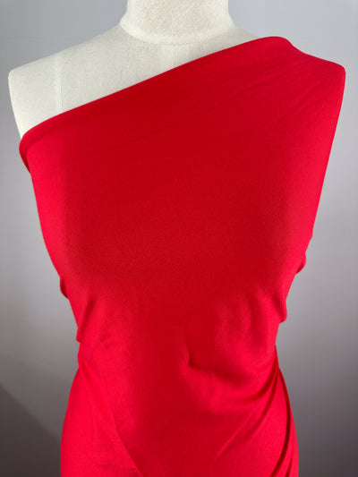 A mannequin displays the Super Cheap Fabrics Cotton Jersey in Ribbon Red, highlighting an asymmetrical one-shoulder design. The 165cm medium-weight jersey is smooth with slight stretch, emphasizing its vivid color and rich texture.