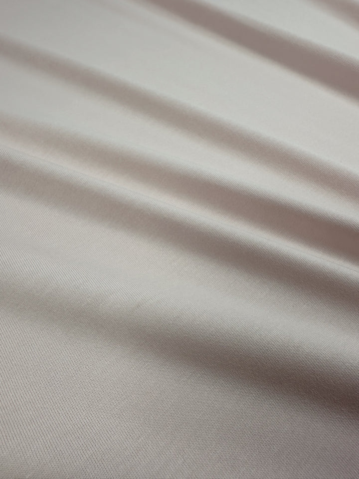 Close-up of Super Cheap Fabrics' Cotton Jersey in Rosewater, featuring subtle diagonal lines and soft folds on a light gray hue. The smooth, slightly reflective texture suggests a silky material ideal for versatile clothing. Width: 165cm.