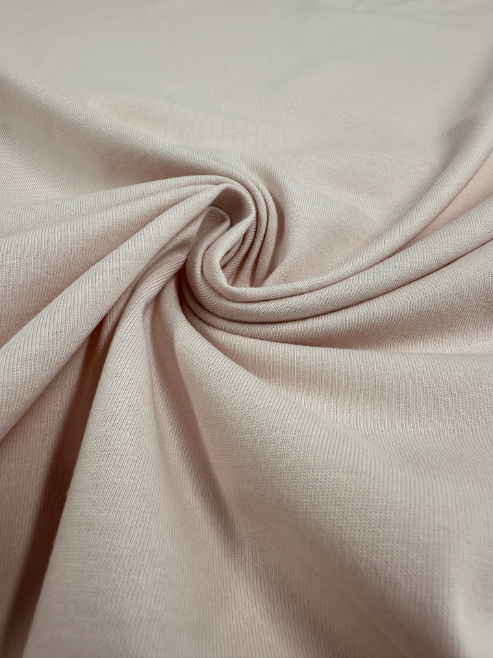 A close-up of Super Cheap Fabrics' Rosewater Cotton Jersey (165cm) showcases its soft, light beige texture and subtle weave. Smooth and lightweight, the fabric’s gentle folds create shadows and highlights, making it ideal for versatile clothing creation.