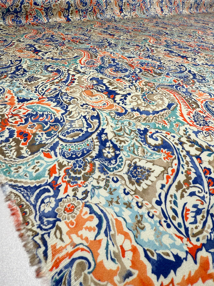 A close-up of Super Cheap Fabrics’ Mercerised Rayon - Paisley Fusion (150cm), showcasing a lightweight rayon textile with swirling blue, orange, beige, and brown paisley patterns and intricate floral and leaf motifs.