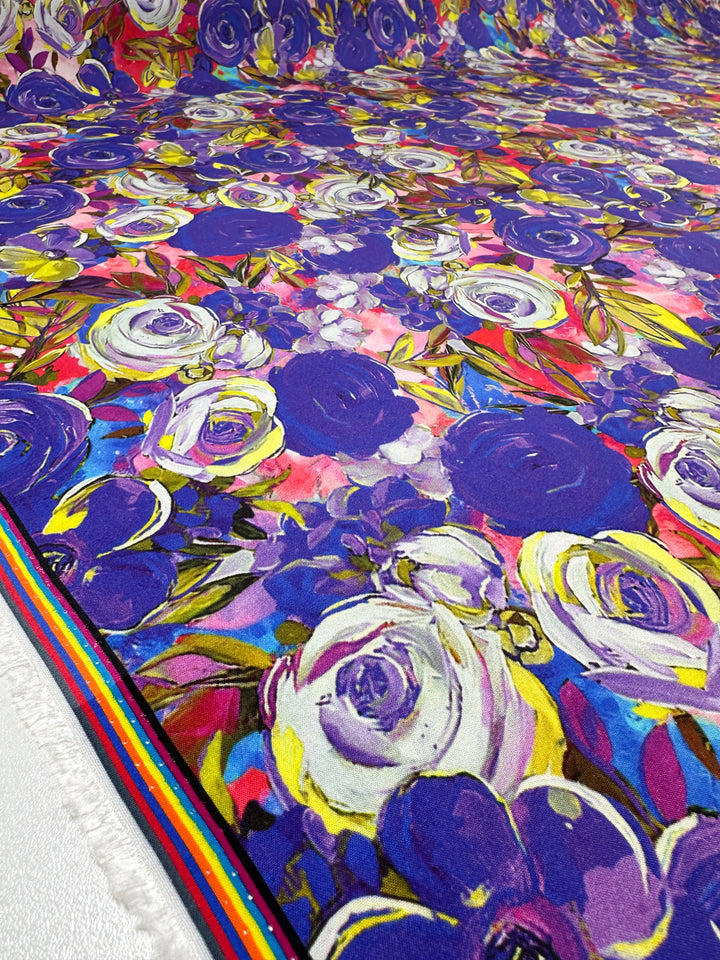 The Zoe Designer Rayon fabric by Super Cheap Fabrics offers a vibrant floral pattern with blue, purple, pink, and white blooms and leaves. It features a colorful striped edge on the left against an abstract backdrop. Width: 145cm.