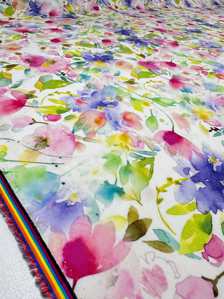 The Designer Rayon - Milana - 145cm by Super Cheap Fabrics showcases a colorful floral watercolor pattern with pink, purple, blue, and yellow flowers on white rayon fabric. A rainbow stripe design edges the fabric, adding versatility to any project.