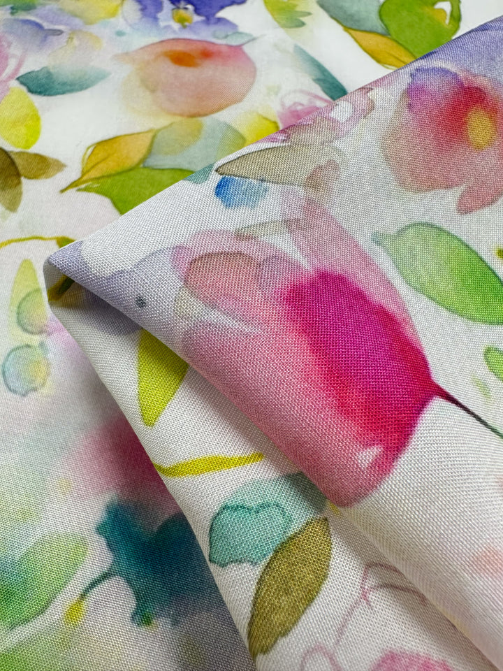 A vibrant floral print on the Designer Rayon - Milana - 145cm by Super Cheap Fabrics features watercolor-style flowers and leaves in pink, yellow, green, and blue. The folded corner highlights its texture and versatile design.