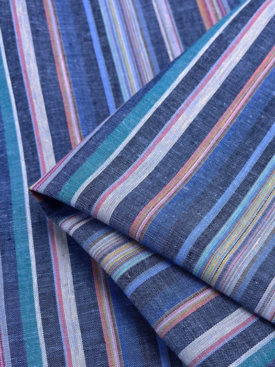 A close-up of the folded, lightweight **Linen Blend - Lunar - 150cm** fabric from **Super Cheap Fabrics** showcases a pattern of multicolored vertical stripes. The yarn-dyed stripes vary in width and include shades of blue, pink, white, and green for a vibrant and textured look.