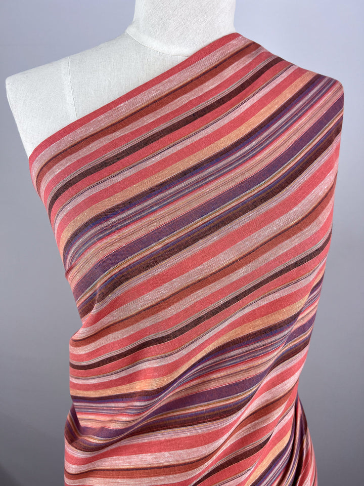 A mannequin is dressed in the Linen Blend - Colombo - 150cm from Super Cheap Fabrics, featuring yarn-dyed fabric with diagonal stripes in shades of red, pink, brown, and purple. This linen fabric offers a vibrant and textured appearance that creates a visually striking vintage look.