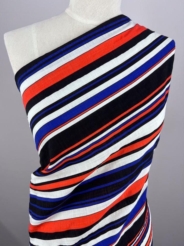 A mannequin displays the "Linen Blend - Minimal - 145cm" from Super Cheap Fabrics, elegantly arranged to cover one shoulder. The breathable, lightweight fabric features a striking pattern of bold diagonal stripes in red, white, black, and blue.